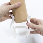 Dos manos colocan una lata impresa en 3D con material tipo madera sobre un soporte blanco, también impreso en 3D, diseñado para sujetar botellas o latas en una silla de ruedas.
