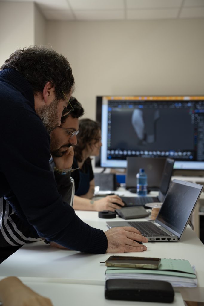 Persona enseñando algo en un ordenador a otra persona con la mano apoyada en el rostro, en un entorno de formación con otras personas al fondo.