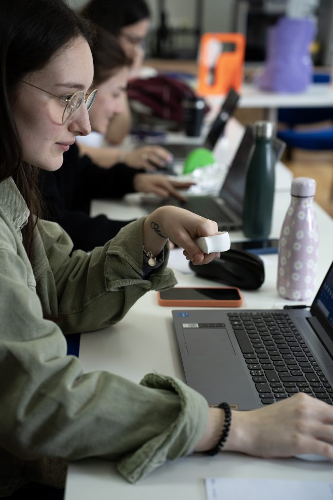 Persona con gafas trabajando con un ordenador portátil mientras sujeta una pieza blanca impresa en 3D en la mano, en una mesa con otros dispositivos y objetos.
