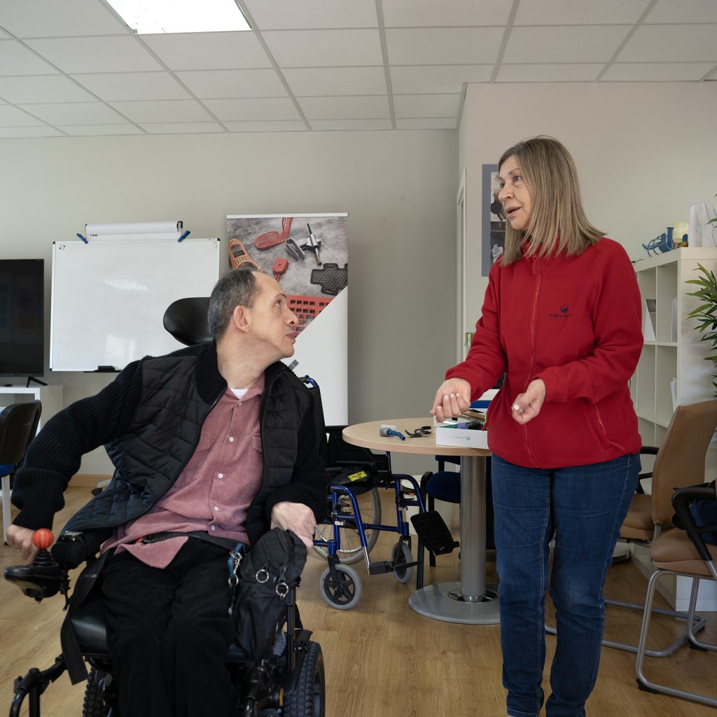 Persona en silla de ruedas conversando con otra persona de pie en un espacio de trabajo con mesas y material técnico.