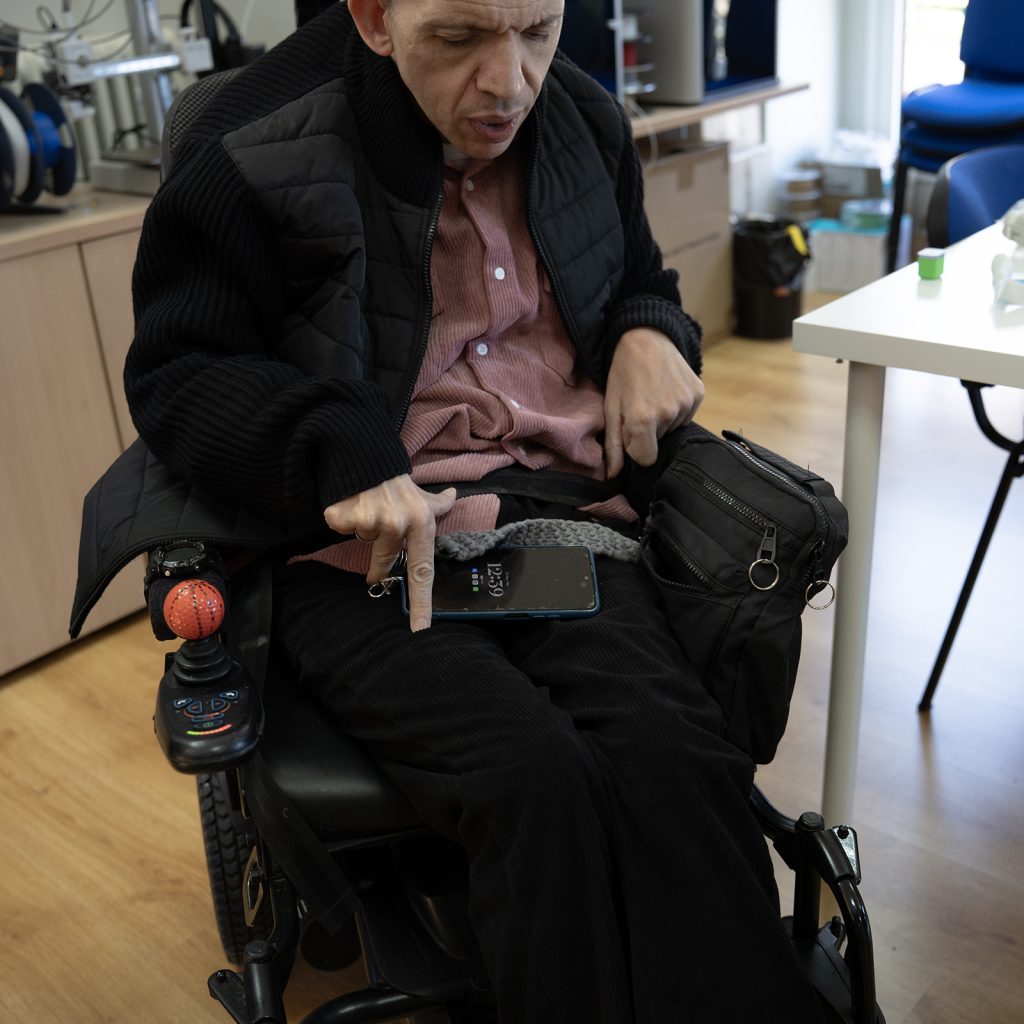 Persona en silla de ruedas señalando un teléfono móvil colocado sobre sus piernas.
