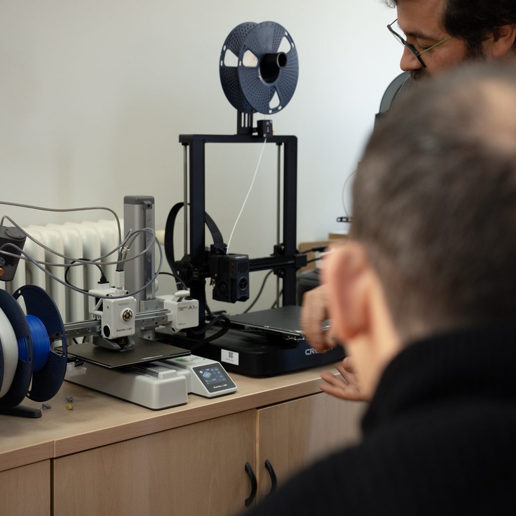 Varias impresoras 3D sobre una mesa con bobinas de filamento, mientras dos personas observan su funcionamiento desde el frente.