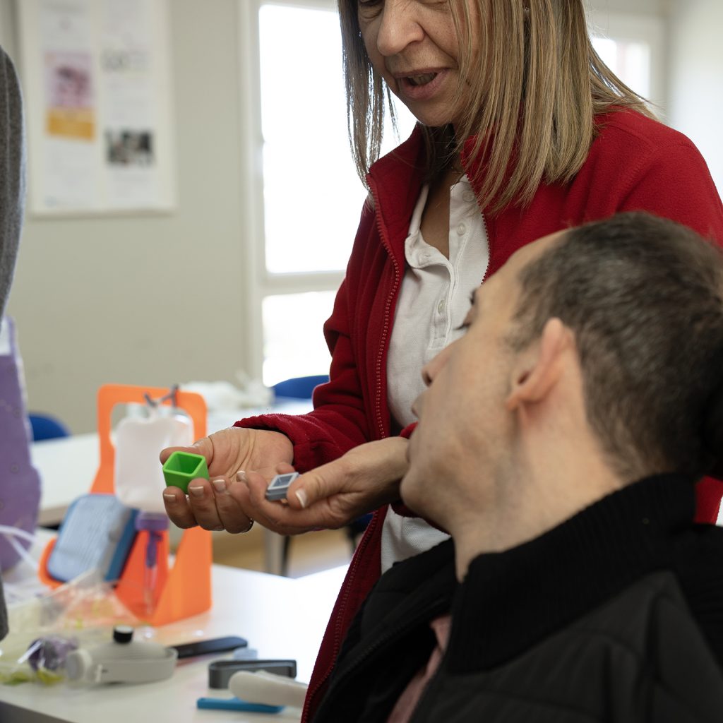 Persona mostrando pequeñas piezas mientras otra persona en silla de ruedas observa, con objetos y productos de apoyo sobre la mesa.