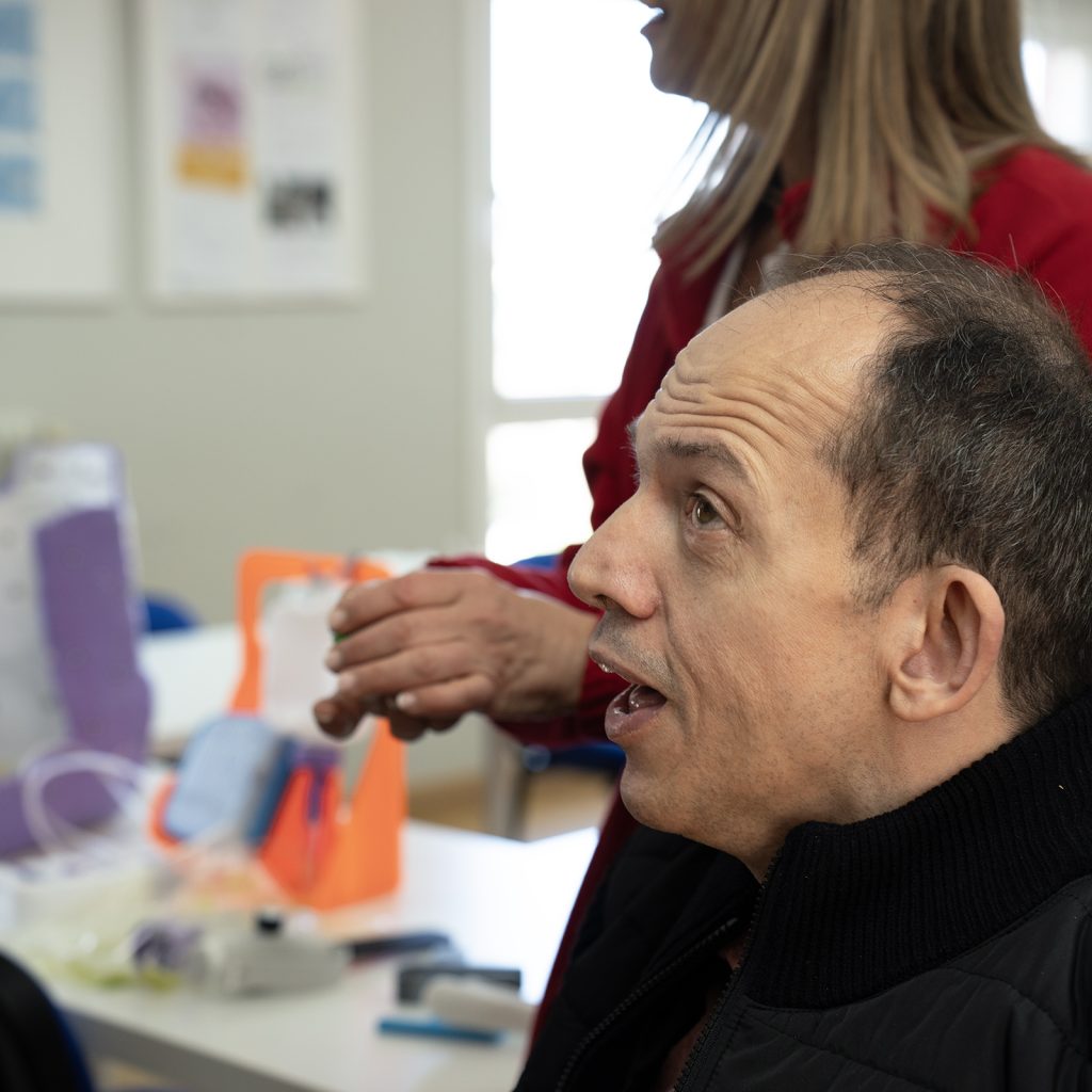 Primer plano de una persona en silla de ruedas mirando hacia otra persona que sostiene un objeto, con material de trabajo al fondo.