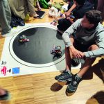 Estudiantes y profesores rodean un tatami circular donde dos robots minisumo compiten entre sí durante el evento CantabRobots + Math 2026.