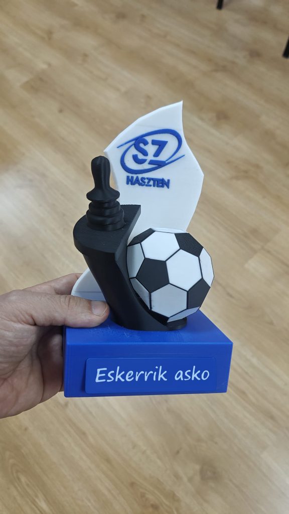 Mano sosteniendo un reconocimiento impreso en 3D con un balón de fútbol, un joystick de mando de silla de ruedas, el logotipo de Haszten y el texto "Eskerrik asko".